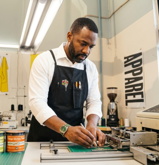 Artisan wearing an apron and mask, precisely measuring and cutting wood in a workshop setting.